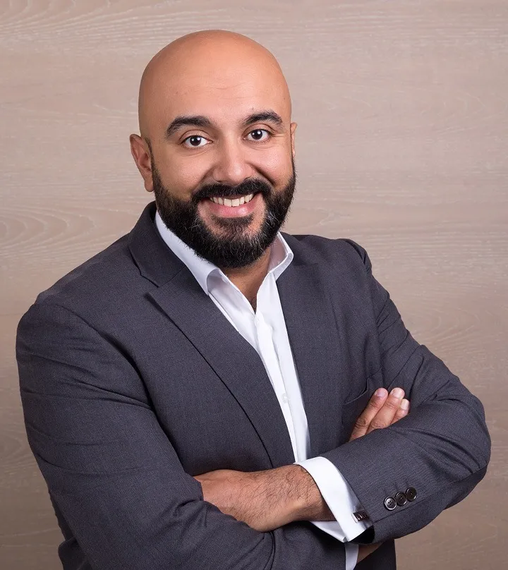 A confident, smiling man with a beard, wearing a dark suit and white shirt, arms crossed, representing leadership and success as discussed on JOH Partners' podcast with Peppy Dosanjh.