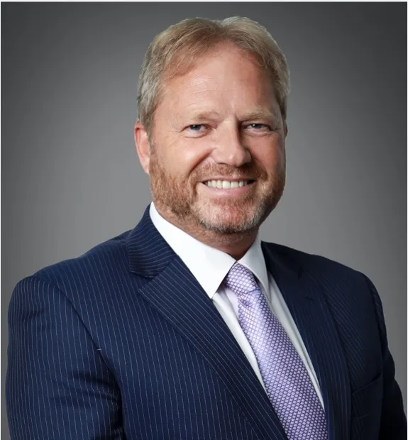 Portrait of Gary Hubbard, a middle-aged Caucasian man with light brown hair, smiling confidently in a navy blue pinstripe suit and light purple tie, representing dedication and leadership on JOH Partners' podcast.