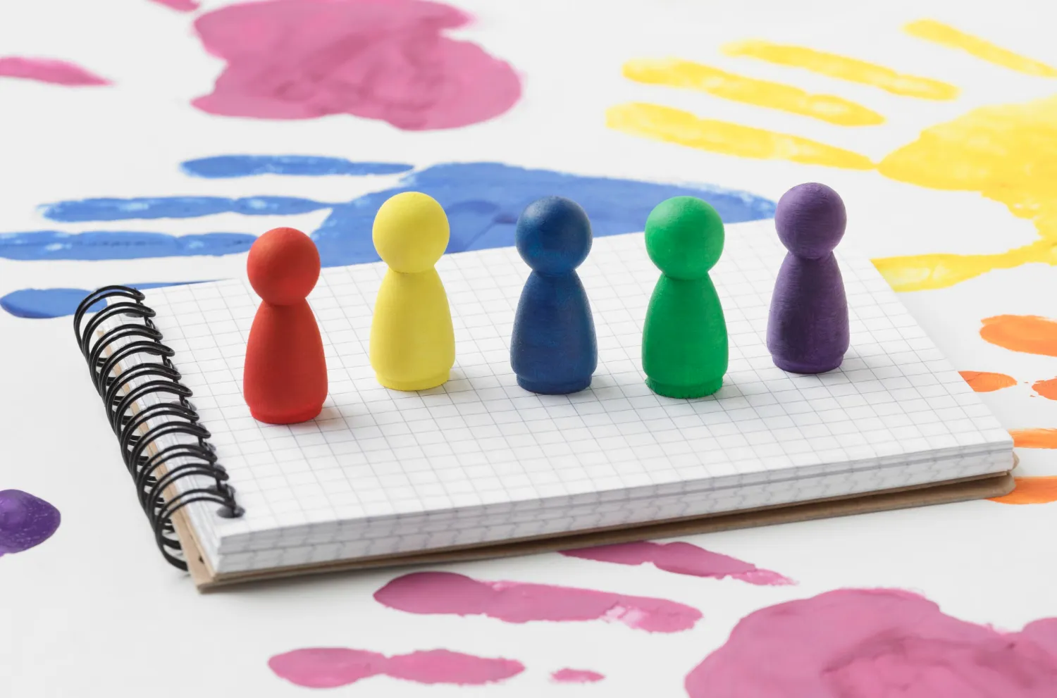 Colorful game pawns on a grid notebook surrounded by multicolored paint splashes, representing the diverse aspects of stakeholder engagement discussed in JOH Partners' blog on Stakeholder Engagement Assessment Matrix.