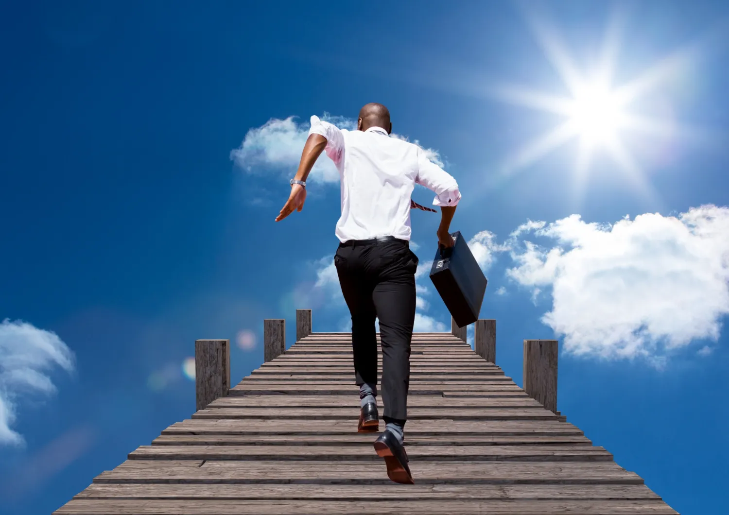 A businessman confidently strides upwards on a wooden staircase against a bright sky, symbolizing career progression from manager to director as discussed in JOH Partners' blog on career advancement.